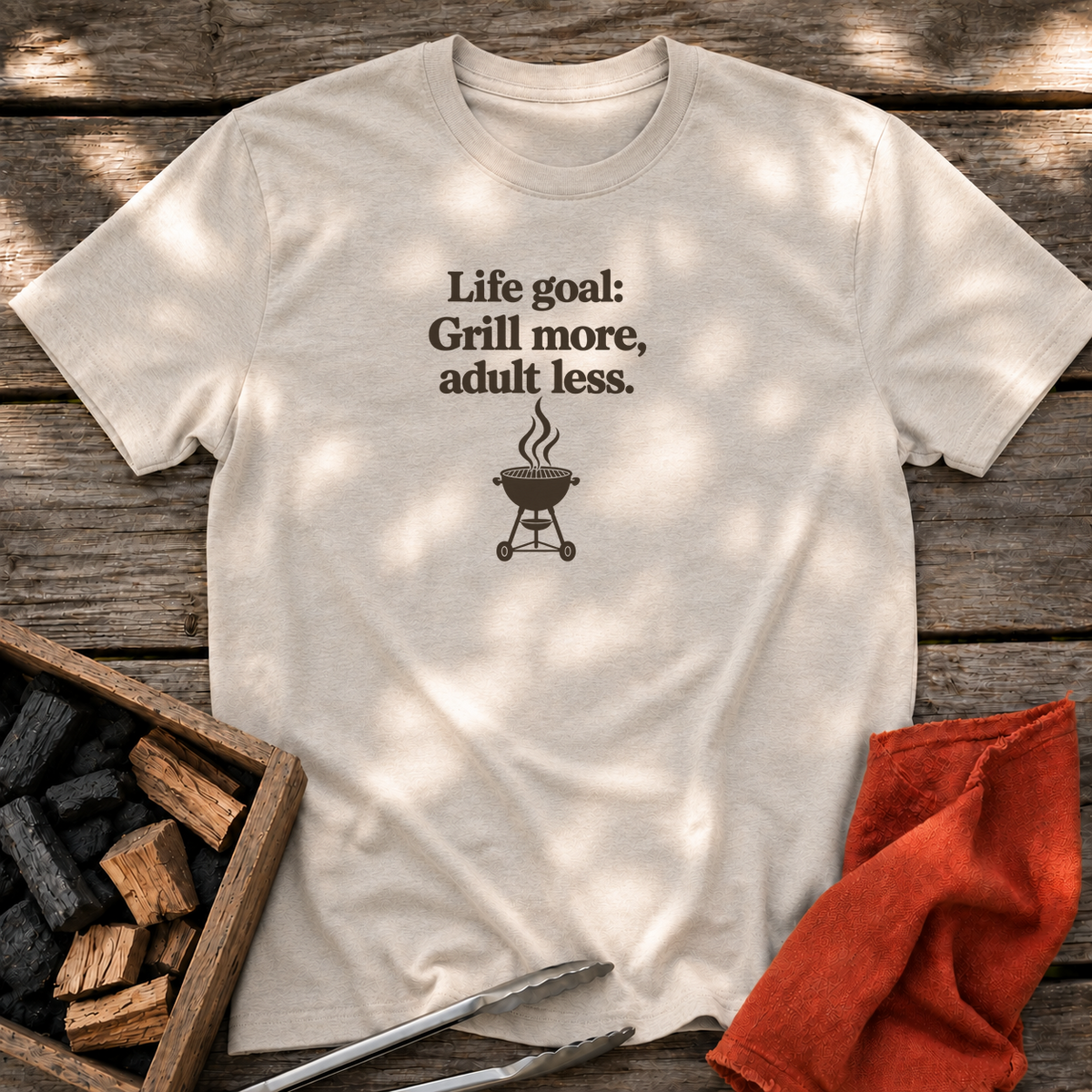 Natural T-shirt with the design “Life goal: Grill more, adult less.” printed on the front.