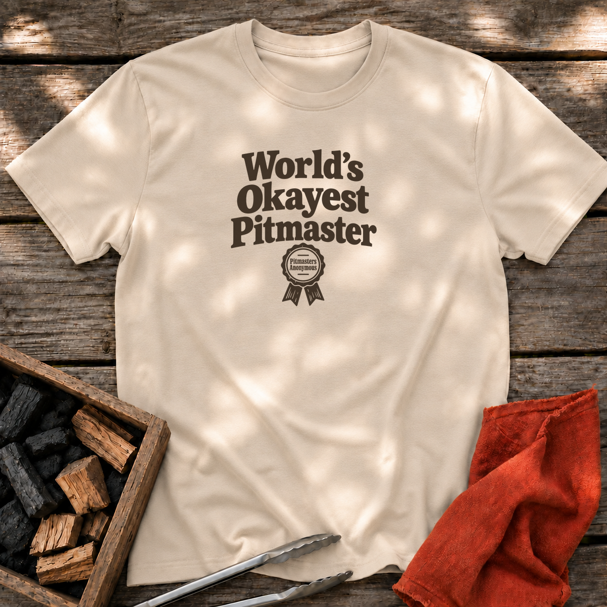 Sand T-shirt with the design “World’s Okayest Pitmaster” printed on the front.