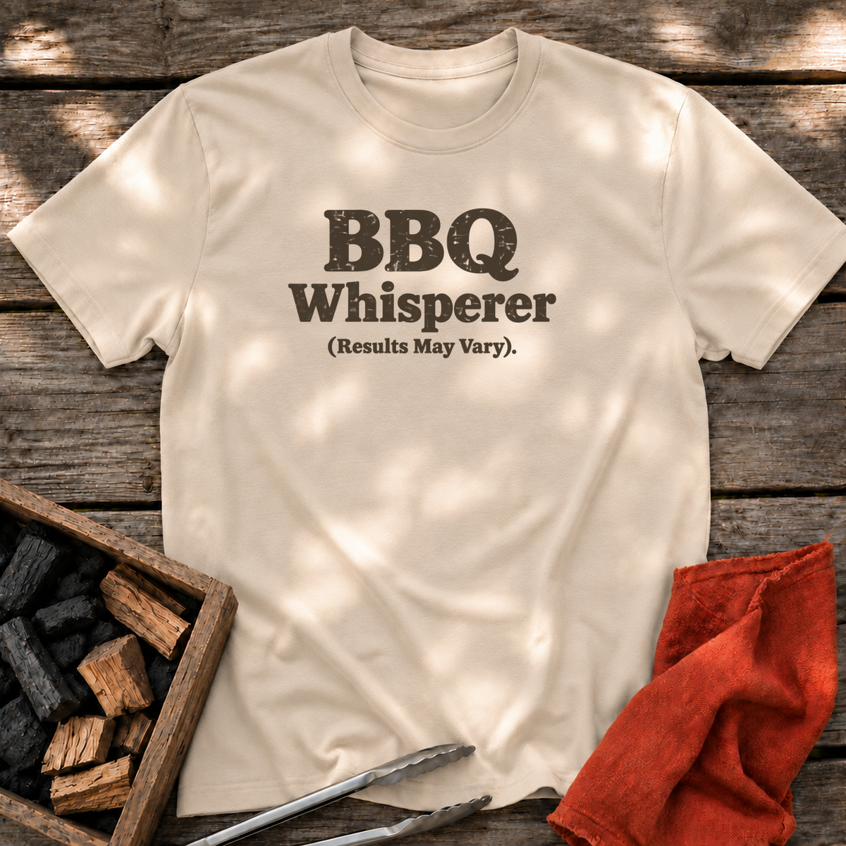 Sand T-shirt with the design “BBQ Whisperer (Results May Vary).” printed on the front.